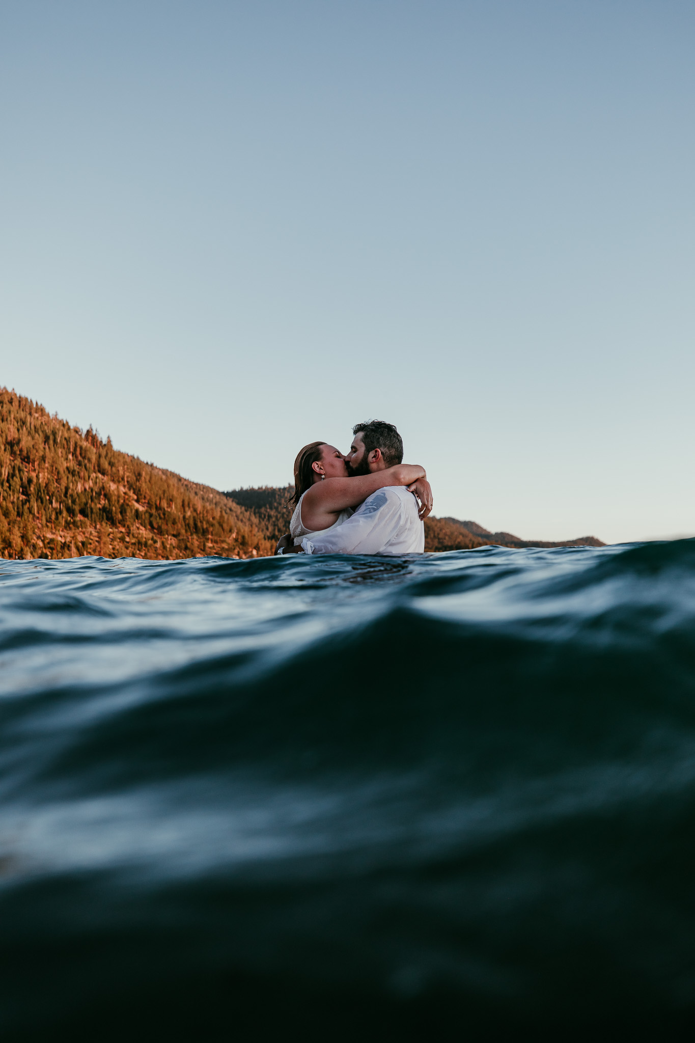 Newlyweds kissing in Lake Tahoe as a wave rises in the foreground at sunset.