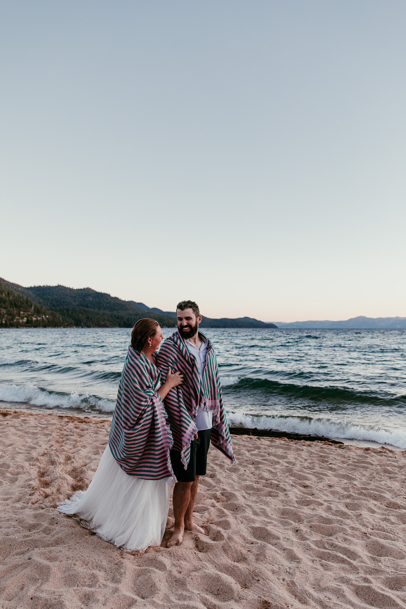 Newlyweds wrapped in towels and leaning together beside Lake Tahoe after their sunset swim.